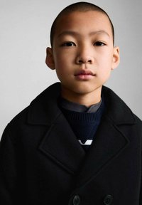 Svart dubbelknäppt kappa med spettslag, bärs över en mörk stickad tröja med vita ränder, med slät textur och minimalistisk design.