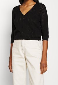 Gilet noir en maille avec un col en V, des manches trois-quarts et un motif en chevron. Fini avec une rangée de boutons et un ourlet côtelé.