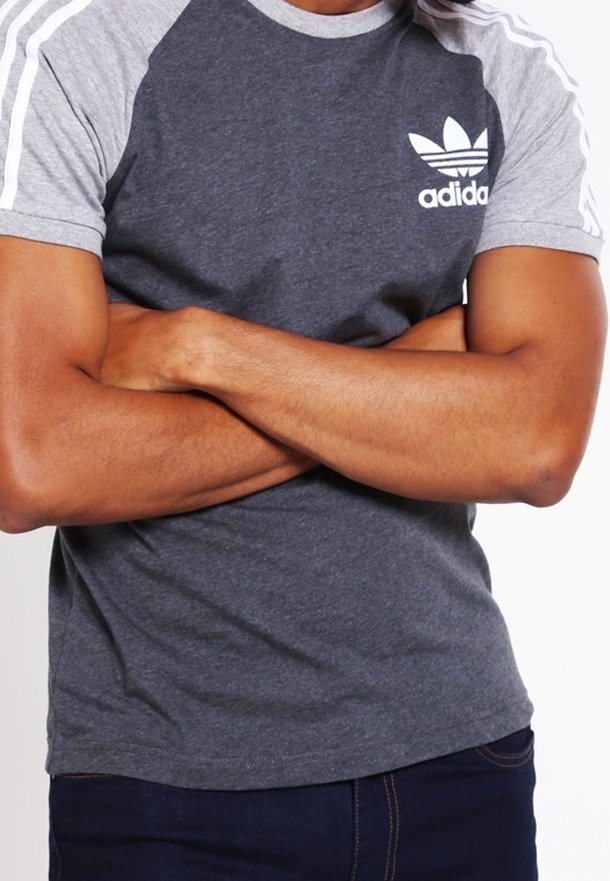 Persona con los brazos cruzados, vistiendo una camiseta de Adidas en gris y gris oscuro con rayas blancas en las mangas y jeans azul oscuro.