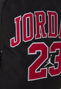 Zaino in tessuto nero con ricamo rosso "JORDAN 23" e logo Jumpman grigio. Il testo ha un bordo bianco per contrasto.