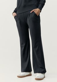 Čierne široké nohavice z elastickej tkaniny s dvoma prednými vreckami a hladkou textúrou. Spárované s bielymi a sivými teniskami.