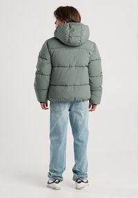 Persona che indossa una giacca imbottita verde con cappuccio, jeans azzurri e sneakers bianche Nike, in piedi di spalle su uno sfondo semplice.