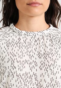 Blusa bianca con una trama testurizzata di piccole forme marroni, scollatura rotonda, tessuto liscio e una leggera lucentezza.