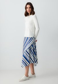 Haut à manches longues blanc associé à une jupe portefeuille bleu et blanche à rayures, avec un design fluide et portée avec des baskets blanches.