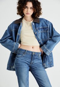 Denim jacket with brown collar, light beige top, and fitted blue jeans. Curly hair adds texture to the overall look.