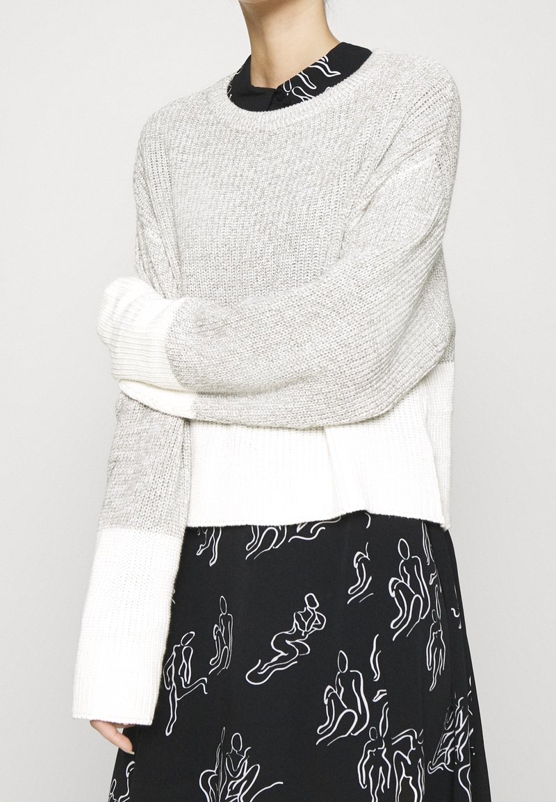 Pull tricoté gris clair et blanc avec texture côtelée, manches longues et ourlet court, porté sur une robe noire ornée de figures abstraites blanches.