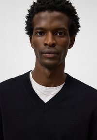 Maglione nero a v-neck realizzato in tessuto a maglia, con una camicia bianca sovrapposta e una semplice collana dorata, caratterizzato da una texture liscia e un logo discreto.
