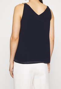 Mörkblå ärmlös topp med V-back, lätt material, har axeldetaljer och rak nederkant. Matchas med vita byxor.