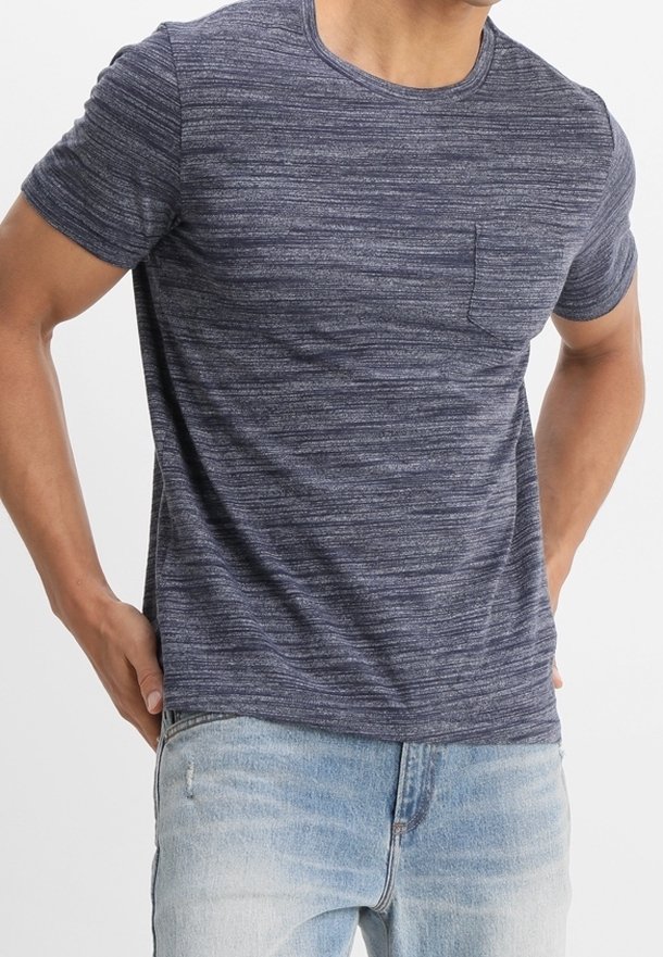 Homme portant un t-shirt ajusté à manches courtes gris anthracite chiné avec une poche poitrine et un jean bleu clair délavé, les mains posées sur les hanches.