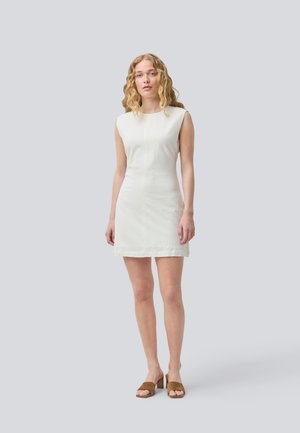 Robe blanche sans manches en tissu texturé avec une silhouette ajustée, atteignant le mi-thigh, associée à des sandales à talons en bloc marron.