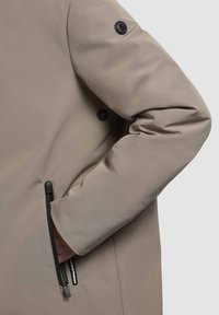 Beige buitenkleding met een gladde textuur, een ritszak en een ronde zwarte logo patch op de mouw. Heeft een getailleerde pasvorm.