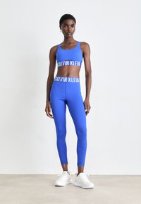 Vrouw staat in een blauwe Calvin Klein sportbeha en legging met witte sneakers tegen een effen witte achtergrond.