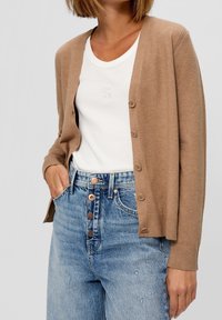 Brun kofta med mjuk textur, knäppning framtill och ribbade muddar, bärs över en vit skjorta och kombineras med ljusblå jeans.