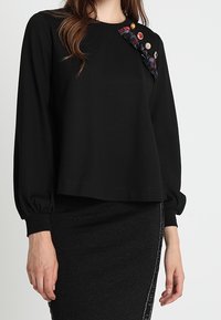 Svart långärmad topp med en avslappnad passform, med en texturerad tyg och en knäppning av färgglada knappar och mönstrat tyg längs axeln.