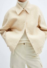 Veste en mélange de laine beige avec un col à revers, fermeture à pression, et épaules tombantes. Portée par-dessus un col roulé côtelé crème et un pantalon clair.