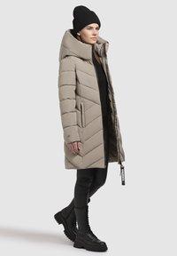 Sieviete, tērpusies bēšā kviltes garā mētelī ar kapuci, melnā cepurē, melnā augšpusē, melnās legingās un melnās šņorētajās zābakos, stāv sānis.