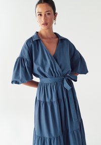 Modrá, vrstvená zavinovací šaty s výstřihem do V, nafouknutými rukávy a páskem v pase. Vyrobeno z lehkého materiálu, který vykazuje hladkou texturu a strukturovaný design.