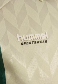 Nærbilde av beige teksturert stoff med geometrisk mønster og "hummel SPORTSWEAR"-logo i hvit og svart skrift.
