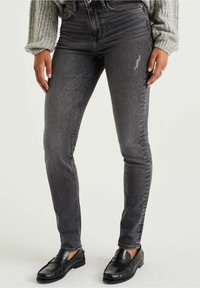 Dunkelgraue Skinny-Jeans mit strukturiertem Finish, hoher Taille, Fünf-Taschen-Design und leichtem Verblassen. Kombiniert mit schwarzen Slippern.
