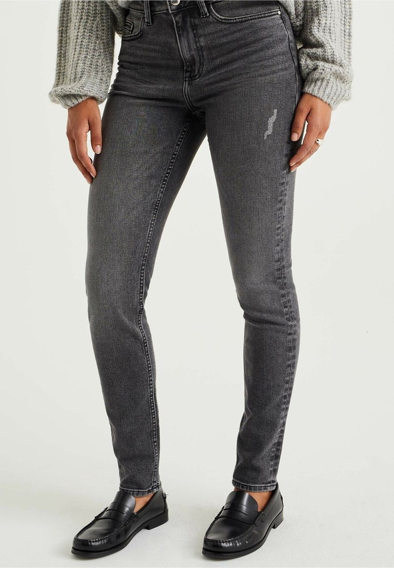 Dunkelgraue Skinny-Jeans mit strukturiertem Finish, hoher Taille, Fünf-Taschen-Design und leichtem Verblassen. Kombiniert mit schwarzen Slippern.