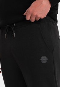 Schwarze Jogginghose mit verstellbarem Kordelzug, die über vordere Taschen und ein hexagonales Logo-Patch auf dem linken Oberschenkel verfügt. Weiches, strukturiertes Material.