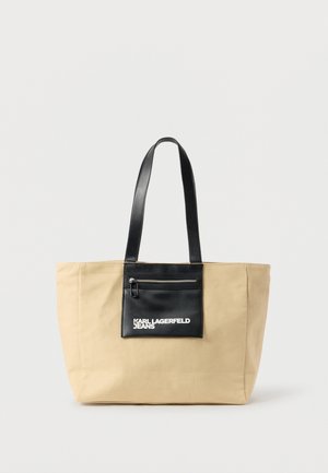 Beige lerretveske med svarte skinnhåndtak og lomme med glidelås foran merket "Karl Lagerfeld Jeans" på hvit bakgrunn.