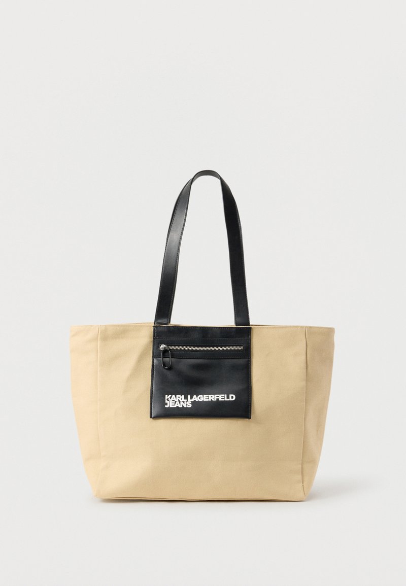 Μπεζ υφασμάτινη τσάντα ώμου με μαύρες δερμάτινες λαβές και μπροστινή τσέπη με φερμουάρ, με την επιγραφή «Karl Lagerfeld Jeans» σε λευκό φόντο.