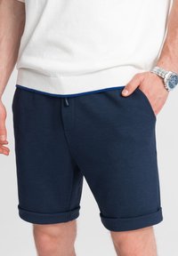 Námořnické modré šortky vyrobené z texturované látky, s páskem na stažení v pase, bočními kapsami a ohrnutými manžetami. Nosí se s bílým svetrem.