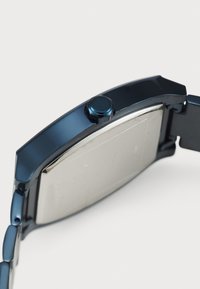 Montre en métal noir avec un boîtier rectangulaire, un dos argenté et une couronne ronde proéminente. Le bracelet présente un design à maillons, soulignant sa durabilité.