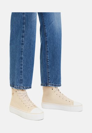 Legs wearing blue jeans and beige high-top sneakers with white thick soles on a plain background.