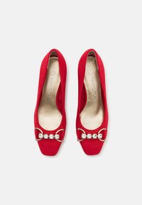 Chaussures à talons hauts en daim rouge avec un embout carré, présentant un accent doré et des bijoux transparents sur la bride avant. Doublure intérieure lisse.