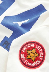 Hvid bomulds T-shirt med et stort blåt grafisk billede og et rundt rødt mærke med teksten "AWESOME EVERYDAY SMILE CHAMPION" og en smilende stjerne.