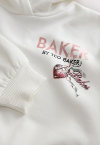 Balta džemperio su gobtuvu su ribotinėmis rankogaliais, ant kurio rausvais rašmenimis parašyta „BAKER“ ir rožinės rankinės grafika su juostele.