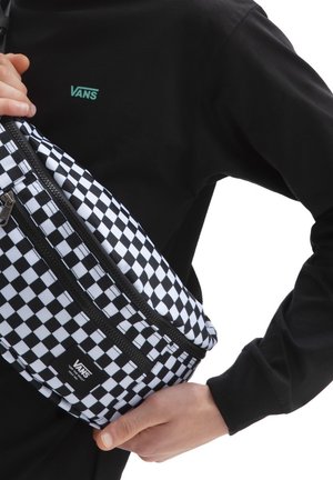 Un sac banane noir et blanc à motif carreaux avec une fermeture éclair, arborant une étiquette avec le logo Vans. Porté en travers du corps sur une chemise noire.