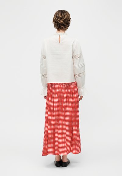 Γυναίκα με κοντά σγουρά μαλλιά, που φοράει λευκό μακρυμάνικο πουκάμισο με δαντελένιες λεπτομέρειες και μακριά κόκκινη φούστα με καρό σχέδιο, στέκεται γυρισμένη προς τα πίσω.