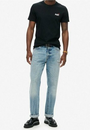 Mężczyzna w czarnej koszulce z krótkim rękawem, jasnoniebieskich dżinsach z podwiniętymi nogawkami, czarnym pasku, czarnych butach typu loafers z frędzlami i złotym zegarku na nadgarstku stoi na tle białym.