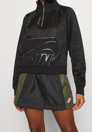 Czarna bluza Nike z zamkiem do ćwiartki, z dużą kieszenią z logo Swoosh i napisem "Just do it.", zestawiona z czarną i oliwkową spódnicą Nike z różowym logo.