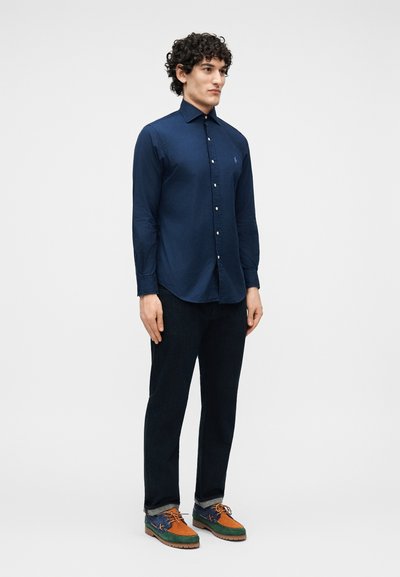 Muž s kučeravými vlasmi, ktorý nosí tmavomodrú košeľu na gombíky, čierne nohavice a viacfarebné hnedé, zelené a modré topánky, stojaci pred jednofarebným pozadím.