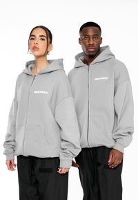 Felpa grigia con zip e tasca frontale, maniche elastiche. Logo nero "MULTIPLY" sul lato sinistro del petto. Indossata con pantaloni neri con patch logo.