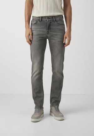 Jeans en denim gris avec un design ajusté, cinq poches et un léger dégradé. Associés à des baskets gris clair. La texture semble lisse avec un éclat minimal.