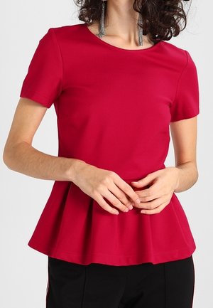 Kobieta ubrana w czerwoną bluzkę peplum z krótkim rękawem i okrągłym dekoltem oraz czarne spodnie, dłonie delikatnie splecione z przodu.