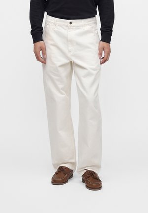 Homme portant un pantalon large blanc cassé, des chaussures bateau marron à lacets, et un haut noir à manches longues, debout devant un fond blanc uni.
