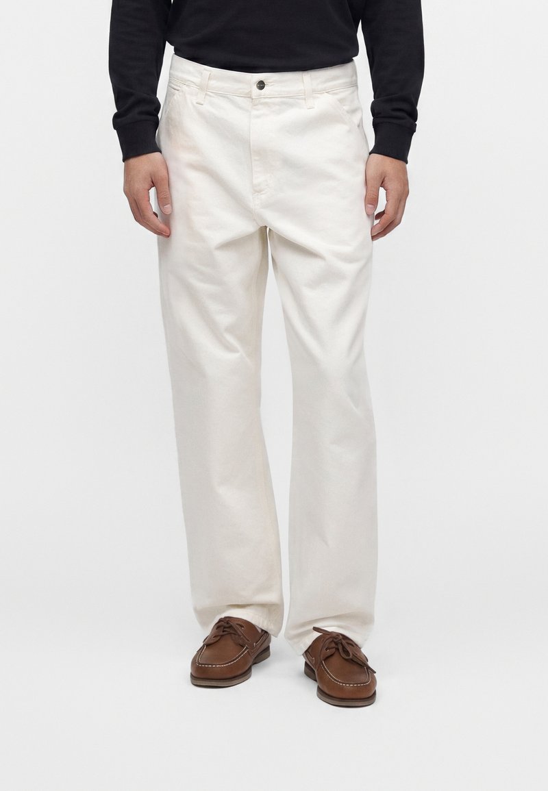 Homme portant un pantalon large blanc cassé, des chaussures bateau marron à lacets, et un haut noir à manches longues, debout devant un fond blanc uni.