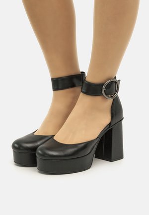 Zapatos de tacón de plataforma negros de cuero con punta redonda, correa en el tobillo y detalle de hebilla circular. El diseño de tacón bloque añade altura.