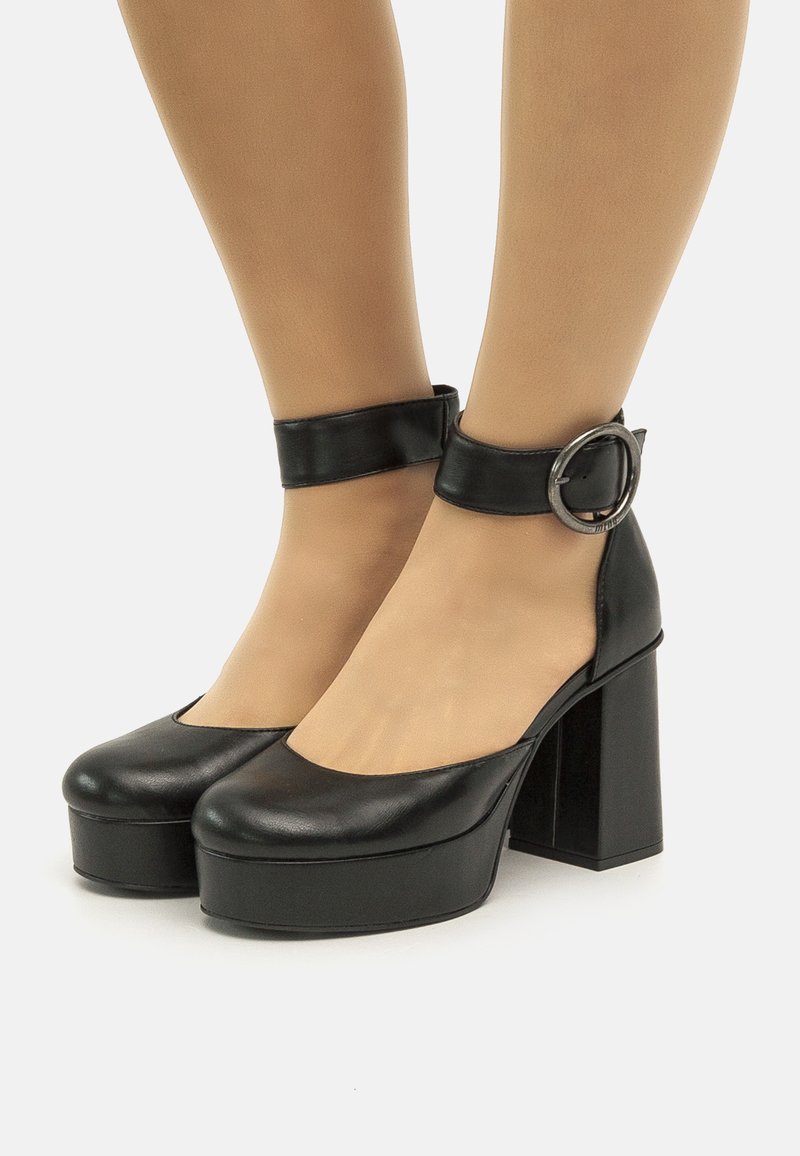 Tacchi a piattaforma in pelle nera con punta arrotondata, cinturino alla caviglia e dettaglio con fibbia circolare. Il design con tacco a blocco aggiunge altezza.