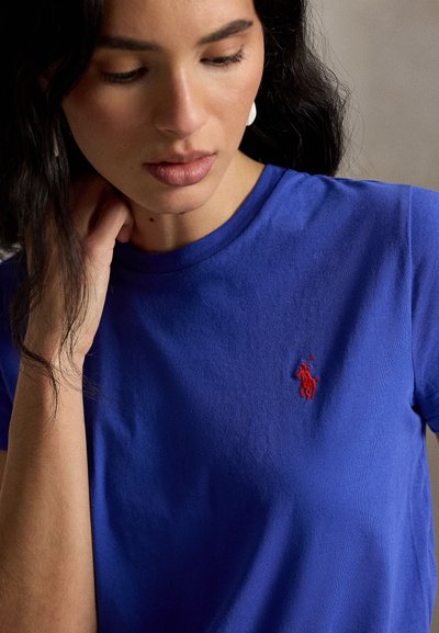 Žena s tmavými vlasmi nosiaca modré tričko s malým červeným vyšívaným logom na hrudi, pozerá sa dolu s rukou blízko krku.