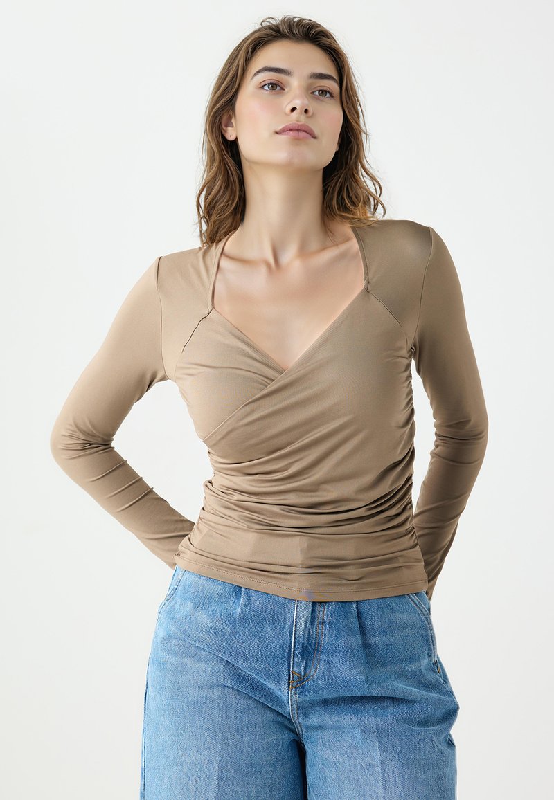 Haut à manches longues marron clair avec un col en V et des détails froncés à la taille, fabriqué en tissu lisse et extensible. Associé à un jean en denim bleu.
