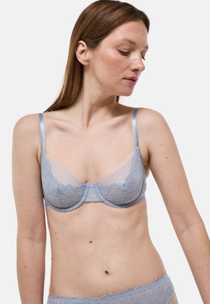 Femme à la peau claire et aux cheveux bruns portant un soutien-gorge en dentelle bleu clair et une culotte assortie, regardant vers le bas à sa gauche sur un fond blanc.