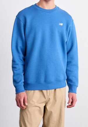 Hombre con sudadera azul lisa de New Balance y pantalones beige, de pie frente a un fondo neutro.