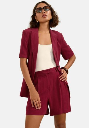 Kvinde iført solbriller, guldringede øreringe, vinrød kortærmet blazer, matchende shorts, hvid top og brun armbånd, poserende med hånden på hoften.